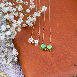 May include: Two gold necklaces with cube pendants. One pendant is white with black letters, the other is green with white letters. The necklaces are on a brown leather notebook, with white flowers in the background.