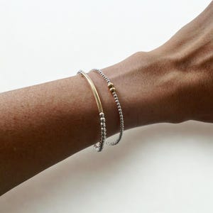 May include: Two bracelets on a wrist. One bracelet is silver with small beads and two gold beads. The other bracelet is silver with small beads and a gold bar. Jewellery.