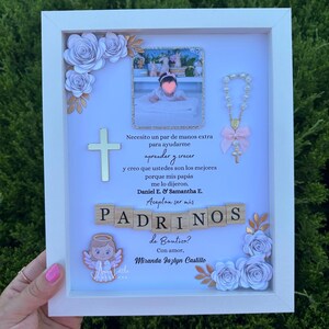 May include: A white framed baptism keepsake with a photo, cross, and decorative flowers. The frame includes a photo of a baby, a cross pendant, and a message in Spanish asking someone to be a godparent.