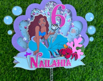 May include: A mermaid-themed cake topper with a mermaid illustration, a number 6, and the name "Nailah" on a banner. The topper features a layered design with glitter accents, bubbles, and a crab.
