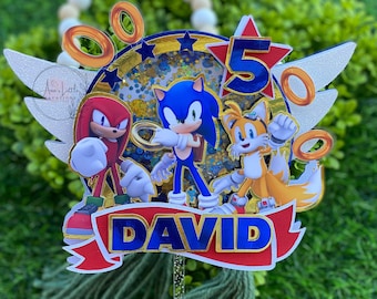 May include: A cake topper featuring Sonic the Hedgehog, Tails, and Knuckles. The topper is decorated with gold glitter, stars, and rings. The name "DAVID" is written in blue and gold letters on a red ribbon.