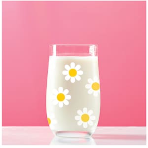 May include: A glass of milk with a white daisy pattern. The glass is filled with milk and has a white daisy pattern on the side. The glass is sitting on a white surface.