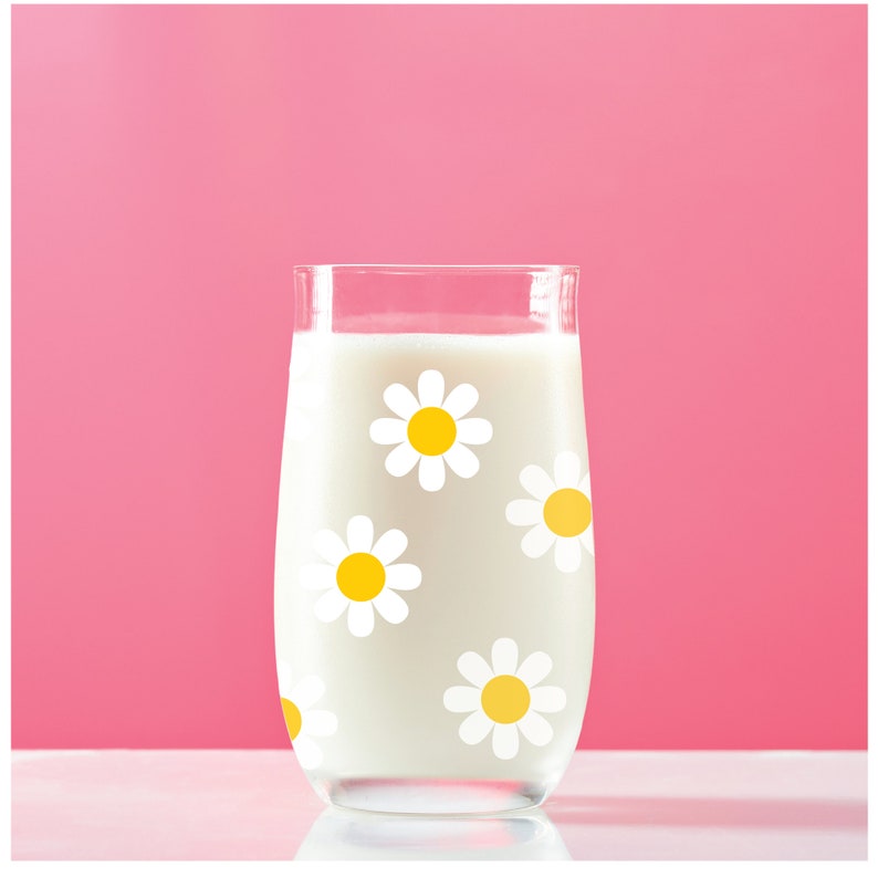 May include: A glass of milk with a white daisy pattern. The glass is filled with milk and has a white daisy pattern on the side. The glass is sitting on a white surface.