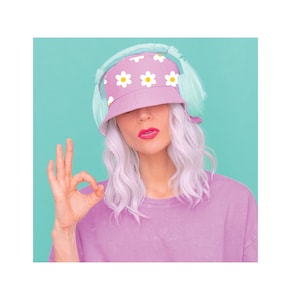 May include: A woman with white hair wearing a pink bucket hat with white daisies and light blue headphones. She is wearing a pink shirt and making an okay sign with her hand.