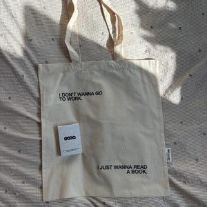 Op de afbeelding: Crèmekleurige draagtas met de tekst "I DON'T WANNA GO TO WORK. I JUST WANNA READ A BOOK." Een kleine witte kaart met de tekst "8000" ligt op de tas. De tas heeft twee crèmekleurige handvatten.
