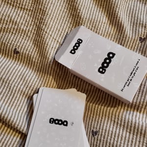 Può includere: Una scatola bianca per giochi di carte e un mazzo di carte con la scritta "8000" stampata sopra. La scatola e le carte sono su un tessuto a righe beige con piccoli motivi a cuore. La scatola ha il testo "54 cartes qui t'aideront à venir à bout de ta pile à lire."