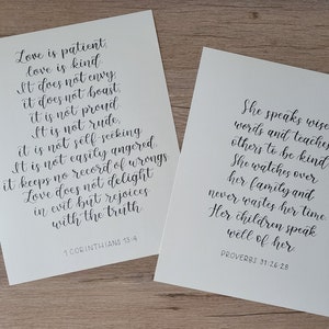 May include: Two white paper prints with elegant calligraphy. The left print features the text "Love is patient, love is kind" from 1 Corinthians 13:4. The right print displays "She speaks wise words" from Proverbs 31:26-28. Both prints have a minimalist design.