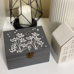 Op de afbeelding: Een grijze houten doos met een wit bloemenmotief en een messing sluiting. De doos staat op een wit oppervlak.