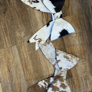May include: Cowhide leather hair bows in black and white, and brown and white patterns. Each bow has a unique pattern and the initials "L/R" are visible on the tips of some bows. The bows are arranged on a wooden surface.