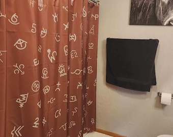 May include: A rust-colored shower curtain featuring a repeating pattern of white western-style cattle brands and symbols. The curtain hangs from a metal rod with circular rings, covering the bathtub area in a bathroom with a dark towel nearby.