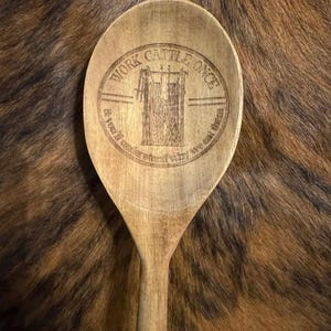 May include: Wooden cooking spoon with an engraved design. The design includes the text "Work Cattle Once & You'll Understand Why We Eat Them" around an image of a cattle gate. The spoon is light brown and has a long handle.