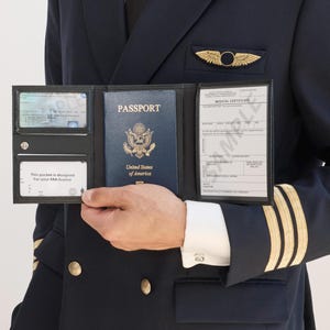 May include: A black leather tri-fold document holder containing a United States passport in the center, a medical certificate on the right, and two clear identification card slots on the left, held by a person in a dark navy blue pilot uniform.