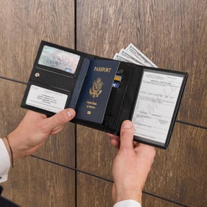 May include: Black leather passport holder with multiple compartments, holding a blue United States passport, cash, and cards. The holder is open, displaying a medical certificate and other documents. The holder is held open by a person.