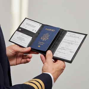 Può includere: Un portadocumenti da pilota in pelle nera a tre ante contenente un passaporto blu degli Stati Uniti al centro. Il lato sinistro presenta due tasche trasparenti con testo stampato per una licenza FAA e un permesso radio. Il lato destro contiene un certificato medico.