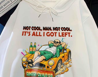 May include: White hoodie with a graphic of a man reclining in a yellow car, text reads "NOT COOL, MAN, NOT COOL. IT'S ALL I GOT LEFT." A black hoodie is next to it. The hoodie has a front pocket and a drawstring hood.