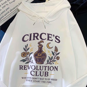 May include: Two hoodies are shown, one white and one black. The white hoodie has a graphic with the text "CIRCE'S REVOLUTION CLUB" and a design of a potion bottle, flowers, and a crescent moon. The text "Witches don't wait to be saved they start the fire" is also present.
