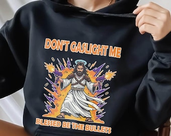 May include: Black hooded sweatshirt with the text "DON'T GASLIGHT ME" above an illustration of a figure holding two guns. Below the illustration is the text "BLESSED BE THE BULLETS". The graphic is in orange, purple, and yellow.