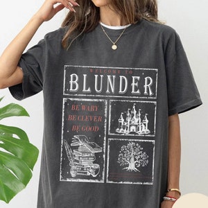 Peut inclure: Un t-shirt surdimensionné gris foncé avec un imprimé graphique qui dit "Bienvenue à Blunder" avec un château, un arbre et des livres. Le texte sous le graphique dit "Soyez prudent, soyez intelligent, soyez bon."