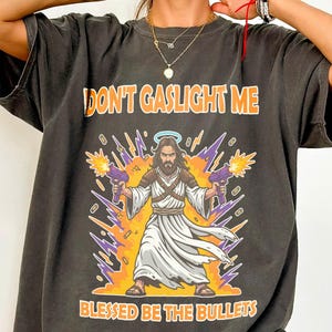 May include: Dark gray t-shirt with the text "DON'T GASLIGHT ME" above an illustration of Jesus holding two purple ray guns. The text "BLESSED BE THE BULLETS" is below the illustration.