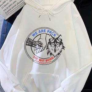 May include: White hoodie with a front pocket and drawstring. The front features a circular graphic with a wolf and the text "WE ARE PACK THE WIT BOND FITZ & NIGHTEYES". A black hoodie is next to it.