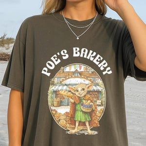 May include: Dark olive green t-shirt with the text "Poe's Bakery" above a circular graphic. The graphic depicts a whimsical creature in a green tunic holding a basket of baked goods in front of a bakery.