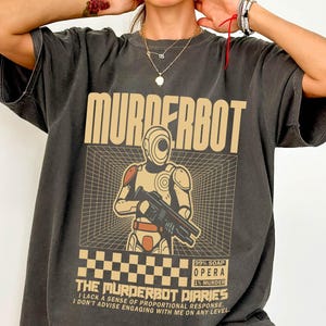 May include: Dark grey t-shirt featuring the word "MURDERBOT" in a tan, blocky font. The shirt has a graphic of a robot holding a gun, with the text "THE MURDERBOT DIARIES". The t-shirt has a loose fit.