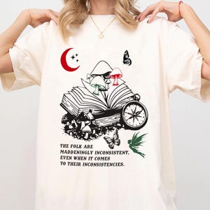 May include: Cream-colored t-shirt featuring a whimsical design with a red crescent moon, mushrooms, butterflies, and a compass. The graphic includes the text: "THE FOLK ARE MADDENINGLY INCONSISTENT, EVEN WHEN IT COMES TO THEIR INCONSISTENCIES."