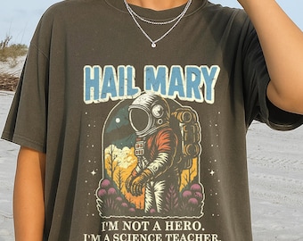 Op de afbeelding: Donker olijfgroen t-shirt met een afbeelding van een astronaut, de tekst "HAIL MARY" erboven en "I'M NOT A HERO. I'M A SCIENCE TEACHER." eronder. De astronaut draagt een ruimtepak met een rugzak, tegen een ruimte- en plantenachtergrond.