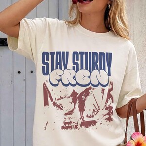 May include: Cream-colored t-shirt with the text "STAY STURDY CREW" in blue and white bubble letters. The shirt features a graphic in burgundy and white. The shirt is short-sleeved.