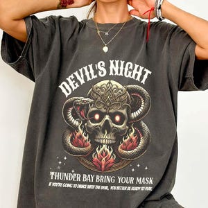 May include: A black oversized t-shirt with a graphic print of a skull with snakes and flames. The text "Devil's Night" is above the skull and "Thunder Bay Bring Your Mask" is below the skull. The text "If you're going to dance with the devil, you better be ready to play" is below the text "Thunder Bay Bring Your Mask".