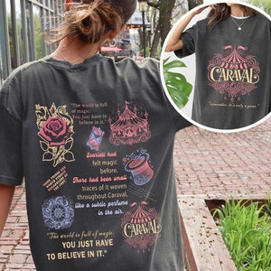 May include: A dark grey t-shirt with a vintage-inspired design featuring a circus tent, a rose, a top hat, and the text "The world is full of magic. You just have to believe in it." and "CARAVAL".