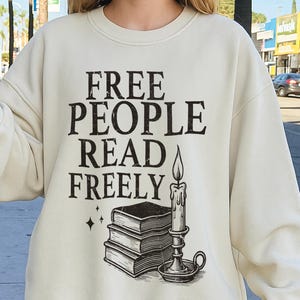 Op de afbeelding: Cr&egrave;mekleurige sweatshirt met de zwarte tekst "FREE PEOPLE READ FREELY". Daaronder een afbeelding van gestapelde boeken, een kaars en sterren. De sweatshirt heeft een casual stijl.