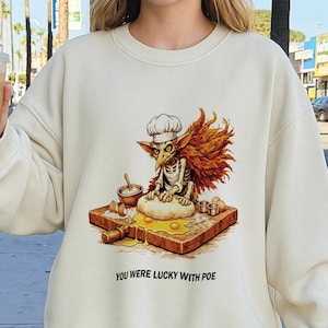 May include: Cream-colored sweatshirt featuring a whimsical illustration of a skeleton chef with a chef's hat, wings, and a baking scene. The text "YOU WERE LUCKY WITH POE" is printed below the graphic.