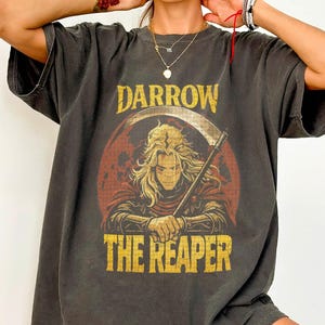 May include: Dark gray t-shirt featuring a graphic of a figure with a scythe, set against a red and orange backdrop. The text "DARROW THE REAPER" is printed above and below the image in a distressed, vintage style.