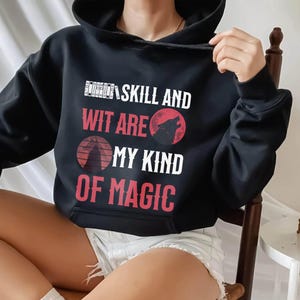 May include: Black hooded sweatshirt with the text "Skill and Wit Are My Kind of Magic" in white and red. The design includes a wolf howling at the moon. The sweatshirt is a casual, comfortable garment.