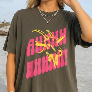 May include: Dark olive green t-shirt with a graphic of a goat head and the text "AHHH HHHH!" in pink and yellow. The shirt is a relaxed fit and has short sleeves.