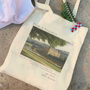 May include: A beige canvas tote bag with a graphic of a building and the text "Pride & Prejudice" and "1813". The quote "There are very few who have heart enough to be really in love without encouragement..." is printed below the graphic. The text "Jane Austen" is printed below the quote.