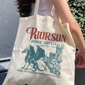 May include: A white canvas tote bag featuring a green dragon and castle graphic. The text on the bag reads "Riorson House Aretia Riders & Ryers Welcome Rebel Faction Basgiath War College".