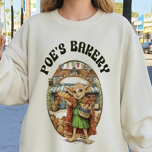 May include: Cream-colored sweatshirt with the text "Poe's Bakery" above an illustration of a cartoon character holding bread and a basket. The character is wearing a green dress and a brown cloak. The bakery has a red roof and a blue awning.