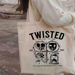 May include: A beige tote bag with black text that reads "Twisted" and "Love - Games - Lies - Bonds". The bag also features a black and white illustration of a broken heart, a mask, a rose, and an hourglass. The text "Est. 2021" is printed on the bag.