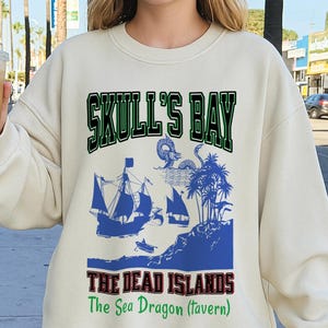 May include: Cream-colored sweatshirt featuring a pirate-themed graphic. The design includes the text "SKULL'S BAY" in green, a blue illustration of ships, islands, and a sea dragon, and the text "THE DEAD ISLANDS The Sea Dragon (tavern)" in red and green.