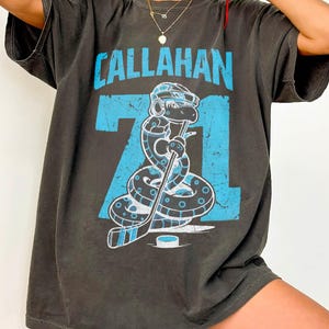 May include: Dark gray t-shirt with the name "CALLAHAN" in light blue block letters above the number "71". The graphic features a snake wearing a hockey helmet and holding a hockey stick. The shirt is oversized.