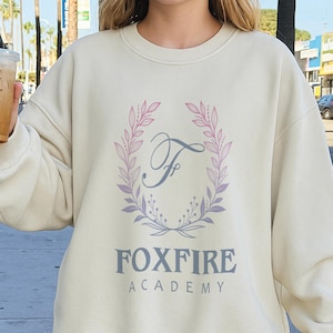 May include: Cream-colored sweatshirt with the text "FOXFIRE ACADEMY" in gray, beneath a floral wreath with the letter "F". The wreath has pink and purple leaves. A casual, long-sleeved top.