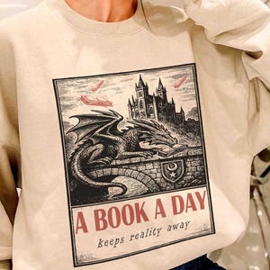 May include: Beige sweatshirt featuring a black and white illustration of a dragon, castle, and the text "A BOOK A DAY keeps reality away." The graphic design is in a vintage style. The sweatshirt is a casual, comfortable garment.