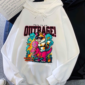 May include: White hoodie with a front graphic of a cat wearing sunglasses riding a pink dinosaur, with the text "THIS IS AN OUTRAGE!". A black hoodie is next to it. The hoodies have a drawstring hood and a front pocket.