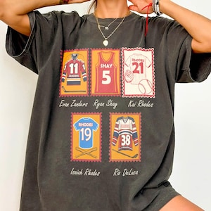 May include: Dark gray t-shirt featuring a stamp-like design with sports jerseys. The jerseys are in various colors, including blue, red, and gold, with numbers and names. The shirt includes the names Evan Zanders, Ryan Shay, Kai Rhodes, Isaiah Rhodes, and Rio Deluca.