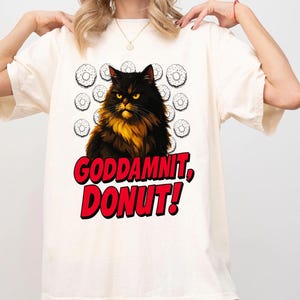 May include: Cream-colored t-shirt featuring a black and gold cat illustration with the text "GODDAMNIT, DONUT!" in red and white. The design includes donut graphics. The shirt is a casual wear item.
