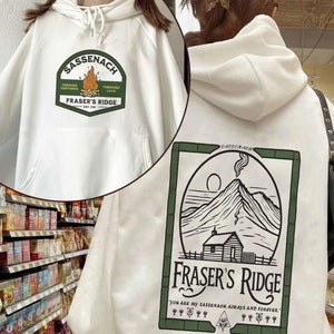 May include: White hooded sweatshirt with a green and black graphic print on the back. The graphic features a mountain range, a cabin, and the text "Fraser's Ridge". The text "You are my Sassenach always and forever." is printed below the graphic.