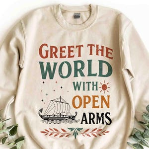 May include: Cream-colored sweatshirt with the text "Greet the World with Open Arms" in a retro font. The design includes a ship, sun, stars, and decorative leaves. A cozy, casual top.