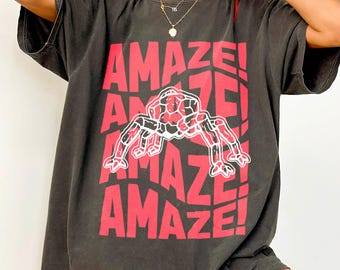Op de afbeelding: Donkergrijs t-shirt met het woord "AMAZE!" in het rood, herhaald. Een rode en zwarte spinnenafbeelding staat in het midden. Het shirt heeft korte mouwen en een losse pasvorm.